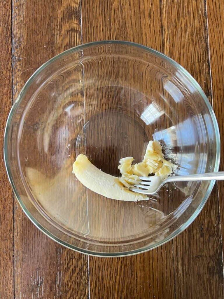 overhead view of a glass bowl with a peeled banana partially mashed by a fork