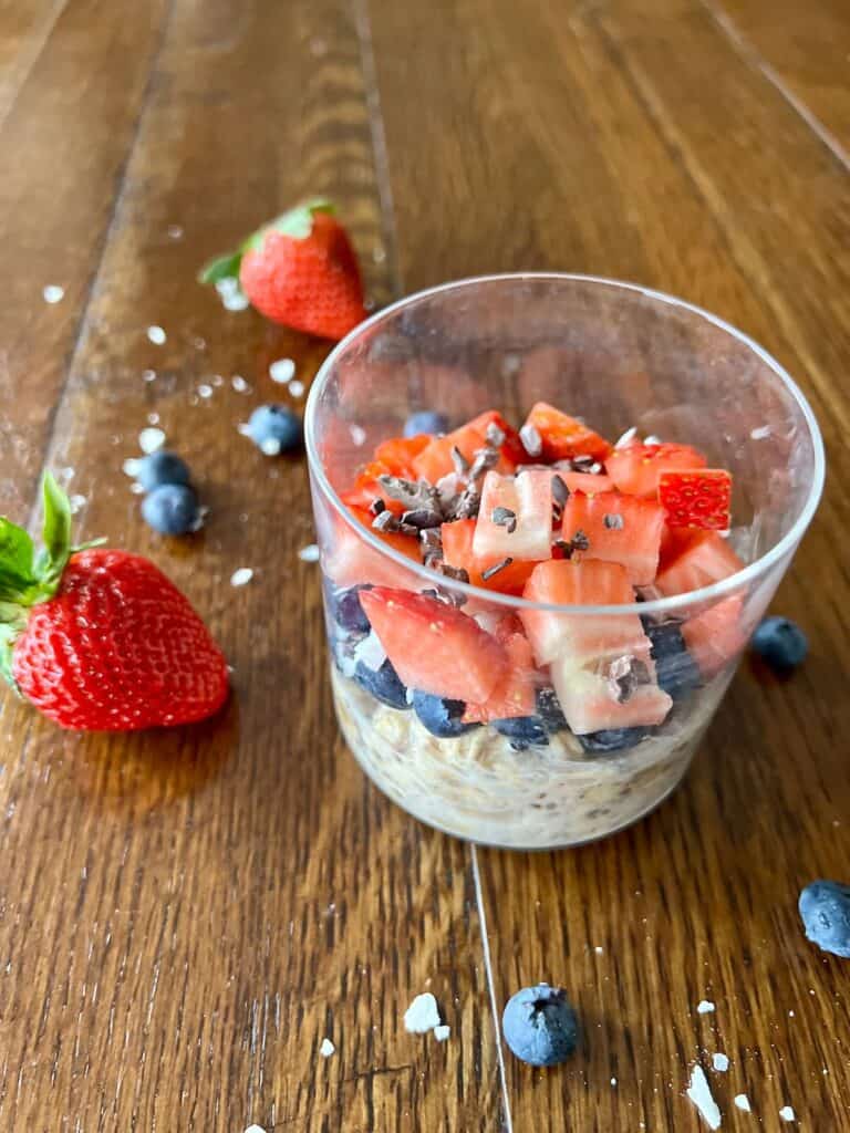 a round glass of classic overnight oats topped with blueberries, strawberries, shredded coconut, and cacao nibs