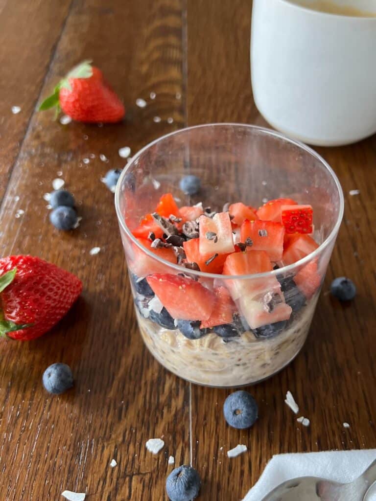 a round glass containing overnight oats topped with blueberries, diced strawberries, shredded coconut, and cacao nibs, with a coffee cup in the background