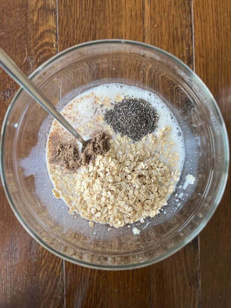 overhead view of a glass bowl containing individually visible ingredients: oats, banana, soy milk, flaxseeds, chia seeds, maple syrup