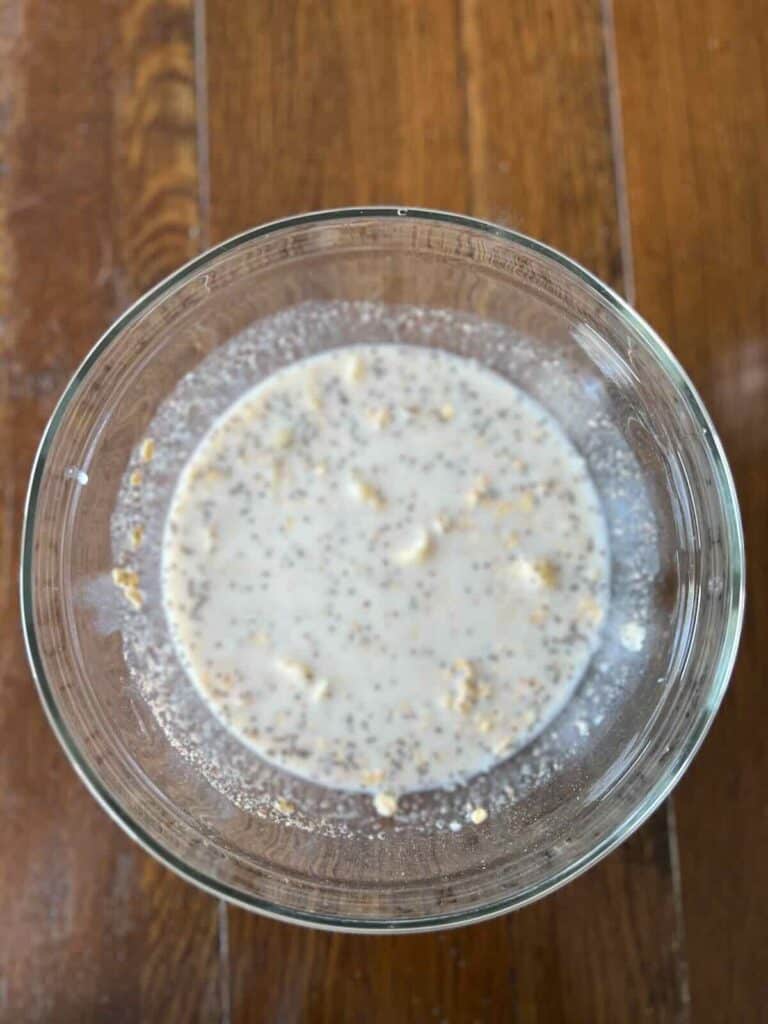 overhead view of a glass bowl containing mixed oats, banana, soy milk, flaxseeds, chia seeds, maple syrup